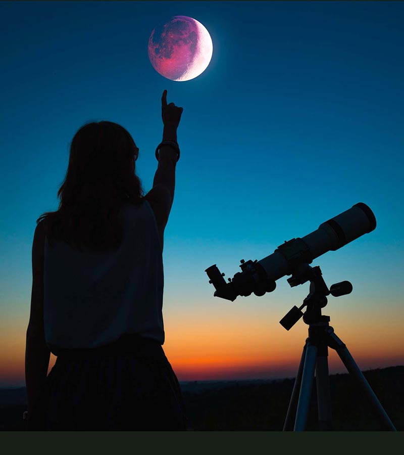 Mujer señalando la luna con el dedo junto a un telescopio. 