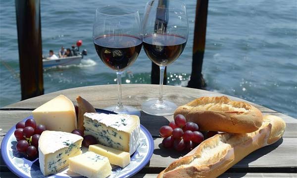 Un plato de queso, pan y uvas en una mesa cerca del agua.